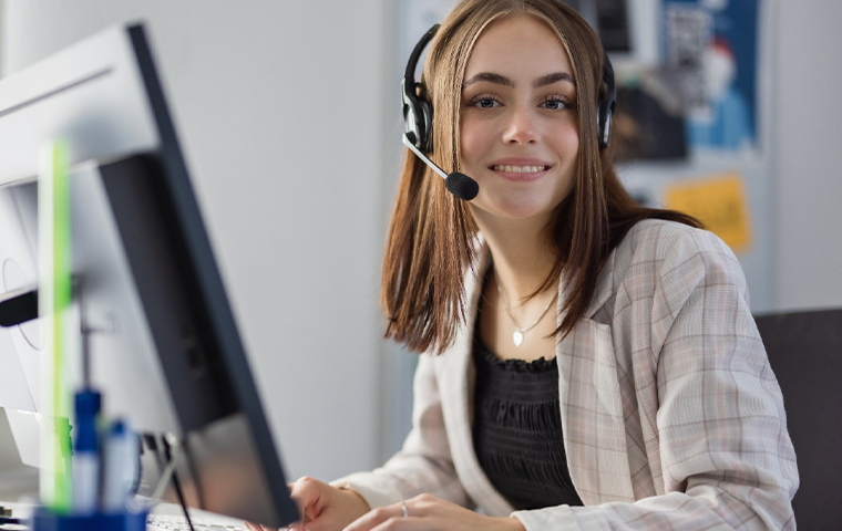 Woman sitting in front of computer facing the camera while wearing a headset. 