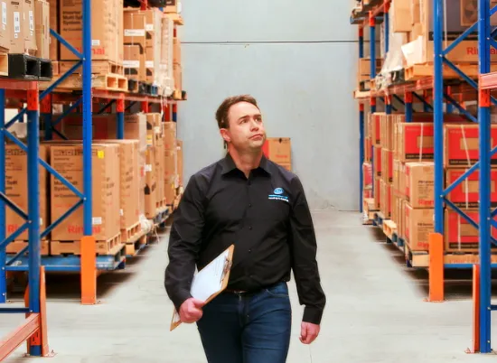 Man holding clipboard and wearing work shirt and jeans walking through warehouse of containers