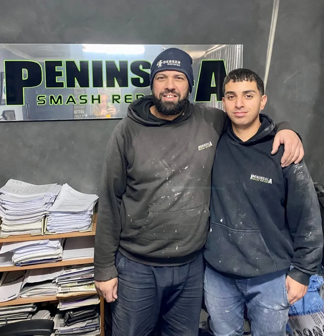 Panel beater standing with work colleague in smash repair shop in front of shop wall decal