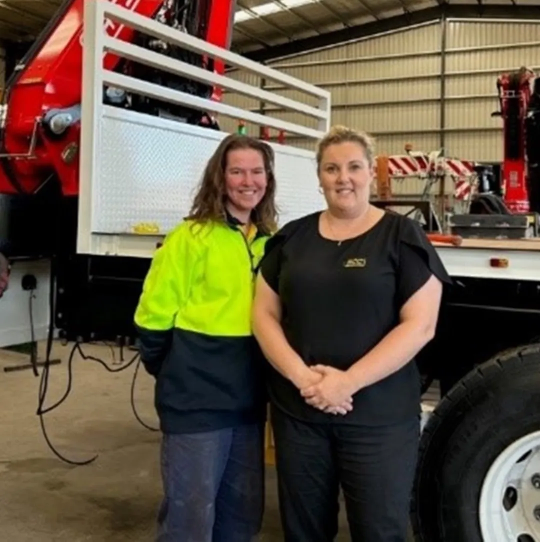 Two women standing in front of a flat bed with crane auto part automobile repair shop