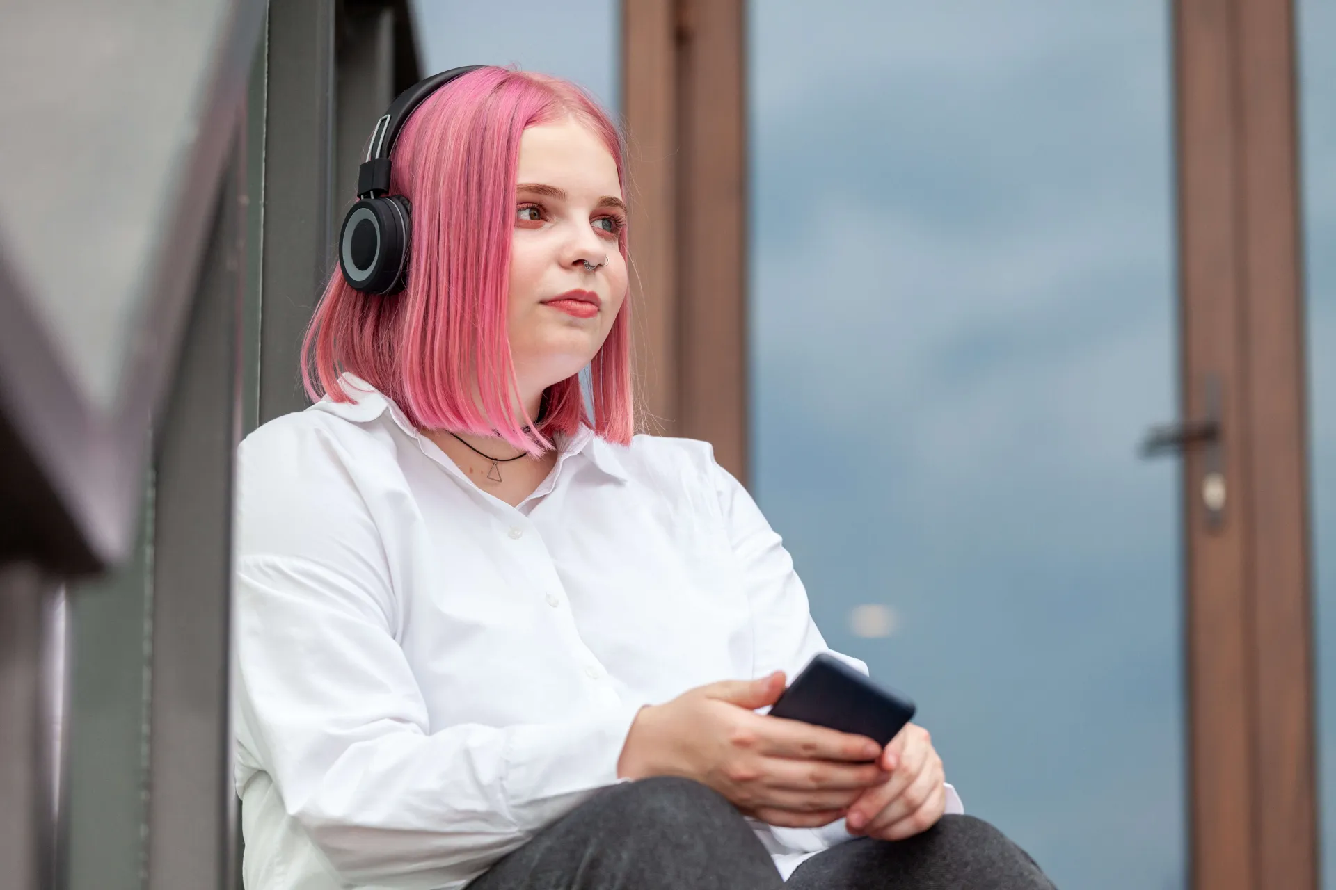 Girl with vibrant pink hair sitting on staircase outside wearing headphones holding a phone.
